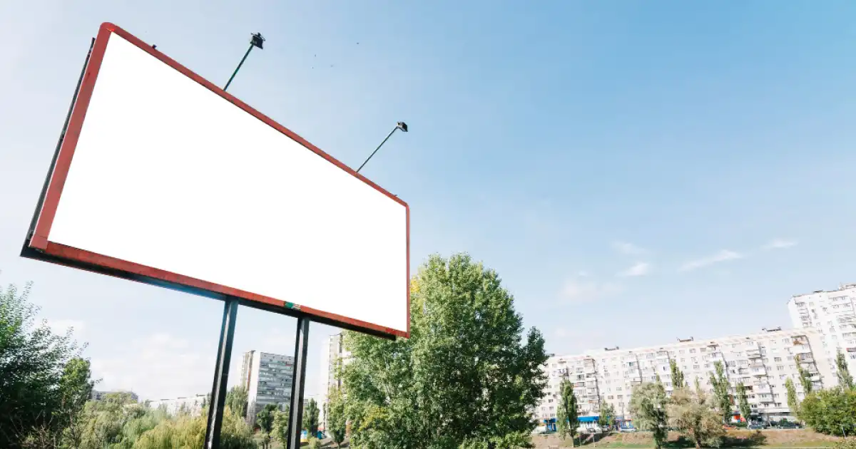 Billboard z pustą powierzchnią reklamową i lampami, na tle drzew oraz zabudowy mieszkalnej