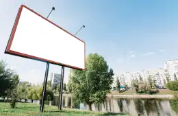 Billboard z pustą powierzchnią reklamową i lampami, na tle drzew oraz zabudowy mieszkalnej